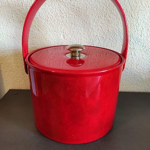Puede incluir: Una cubitera roja brillante con asa y tapa a juego. La cubitera tiene forma cilíndrica y un acabado brillante. La tapa está rematada con un pequeño pomo dorado. Artículo de bar vintage.