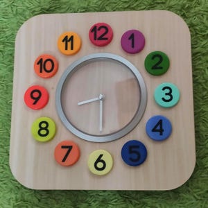 Könnte beinhalten: Pädagogische Holzuhr mit hellem Holzmaserungsfinish. Das Zifferblatt hat eine silberfarbene Lünette und farbige, nummerierte Stundenmarkierungen. Die Zeiger zeigen ungefähr 8:40 Uhr an. Die Zahlen sind in verschiedenen Farben, darunter Rot, Orange und Blau.