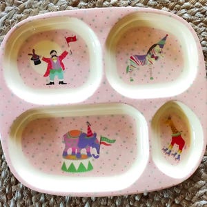 Può includere: Un piatto rosa diviso con illustrazioni a tema circo. Il piatto presenta quattro scomparti ed è decorato con immagini di un presentatore, una zebra, un elefante e una giraffa, tutti in abiti festosi. Il piatto ha un motivo a pois.