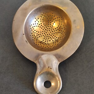 Puede incluir: Un colador de té vintage con un diseño circular y un asa. El colador tiene un cuenco perforado y un acabado plateado. El asa tiene un agujero para colgarlo o guardarlo. El colador de té está diseñado para infusionar té de hojas sueltas.