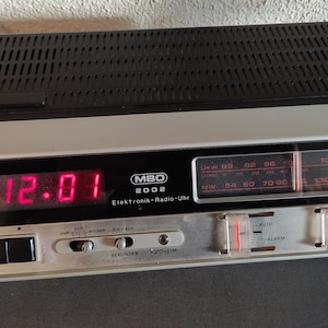 Pode incluir: Um rádio despertador vintage preto e prateado com um visor digital vermelho mostrando 12:01. O rádio tem um dial com marcações de frequência para as bandas UKW e MW. O rádio despertador tem botões para dormir, alarme e atenuamento automático.