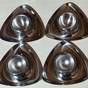Könnte beinhalten: Vier glänzende, silberfarbene Eierbecher. Jeder Becher hat eine dreieckige Form mit einer abgerundeten Mitte, um ein Ei zu halten. Die Becher bestehen aus einem reflektierenden Metall und sind quadratisch angeordnet.