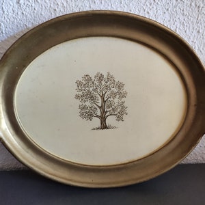 Könnte beinhalten: Ein ovales Serviertablett mit einem goldfarbenen Rahmen und einem weißen Innenraum. Das Tablett zeigt ein schlichtes Design mit einem braunen Baum in der Mitte.