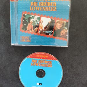 Può includere: Una custodia per CD blu e rossa con il titolo "Die Brüder Löwenherz" e l'autore "Astrid Lindgren". La custodia presenta un'immagine di bambini a cavallo. Il CD è anche visibile, con lo stesso titolo e autore.