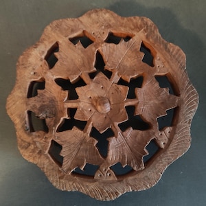 Puede incluir: Un posavasos redondo de madera con un diseño tallado de hojas y flores. El posavasos es marrón y tiene un acabado de madera natural.