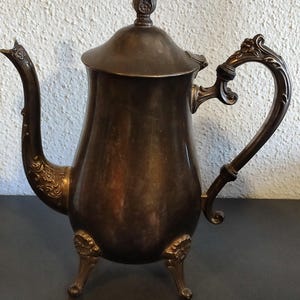 Könnte beinhalten: Eine silberne Teekanne mit einem Blumendesign am Griff und einem dekorativen Knauf auf dem Deckel. Die Teekanne hat drei Beine und einen bauchigen Körper.
