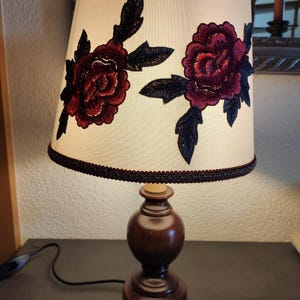 Könnte beinhalten: Eine Tischlampe aus Holz mit einem cremefarbenen Lampenschirm, der mit zwei gestickten roten Rosen mit schwarzen Blättern verziert ist.