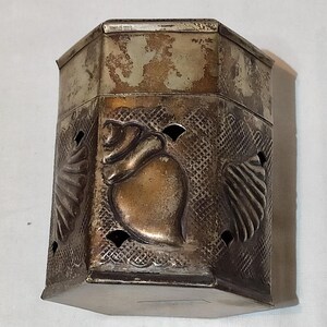 Pode incluir: Uma caixa de metal prateado vintage com formato octogonal. A caixa apresenta desenhos em relevo de flores e corações, com recortes decorativos. O metal tem uma aparência manchada, adicionando ao seu charme antigo. A tampa também é prateada.