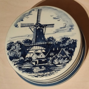 Könnte beinhalten: Eine weiße Keramikdose mit Deckel und einer blauen und weißen Windmühlenszene. Die Windmühle befindet sich im Zentrum der Szene mit Bäumen und einem Fluss im Vordergrund. Der Deckel hat einen blau-weiß gestreiften Rand.