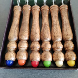 Può includere: Set di sei spiedini di legno con manici marroni e punte colorate. I manici presentano un motivo a venature del legno dettagliato. Le punte sono blu, arancioni, rosse, verdi, bianco sporco e gialle. Gli spiedini sono in una scatola verde scuro.