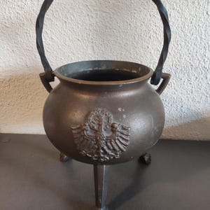 Peut inclure: Un chaudron de couleur bronze avec une poignée torsadée en métal noir et trois pieds. Un emblème en relief est présent sur le devant. Le chaudron a un corps rond et une large ouverture.