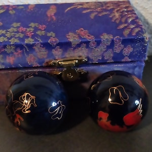 Könnte beinhalten: Eine blaue Schatulle mit goldenem Verschluss und zwei dunkelblauen Zierkugeln. Die Kugeln haben goldene und rote Blumenmuster. Die Schatulle ist blau mit goldenen und rosa Blumenmustern.