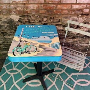 Peut inclure: Une table de bistrot carrée au plateau bleu clair orné d'une illustration graphique de plage, d'un vélo et du texte L'ÉTÉ AUX Îles de la Madeleine Québec Canada, reposant sur un pied central noir, accompagnée d'une chaise en métal bleu clair usé.