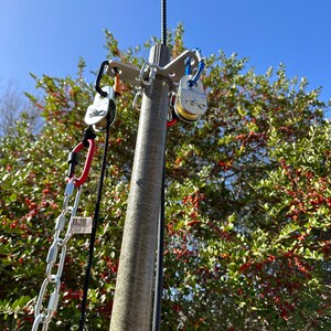 May include: A metal swing set attachment with multiple carabiners and pulleys. The attachment is mounted on a grey pole, with a chain and rope hanging down. The background features a clear blue sky and a tree with red berries.