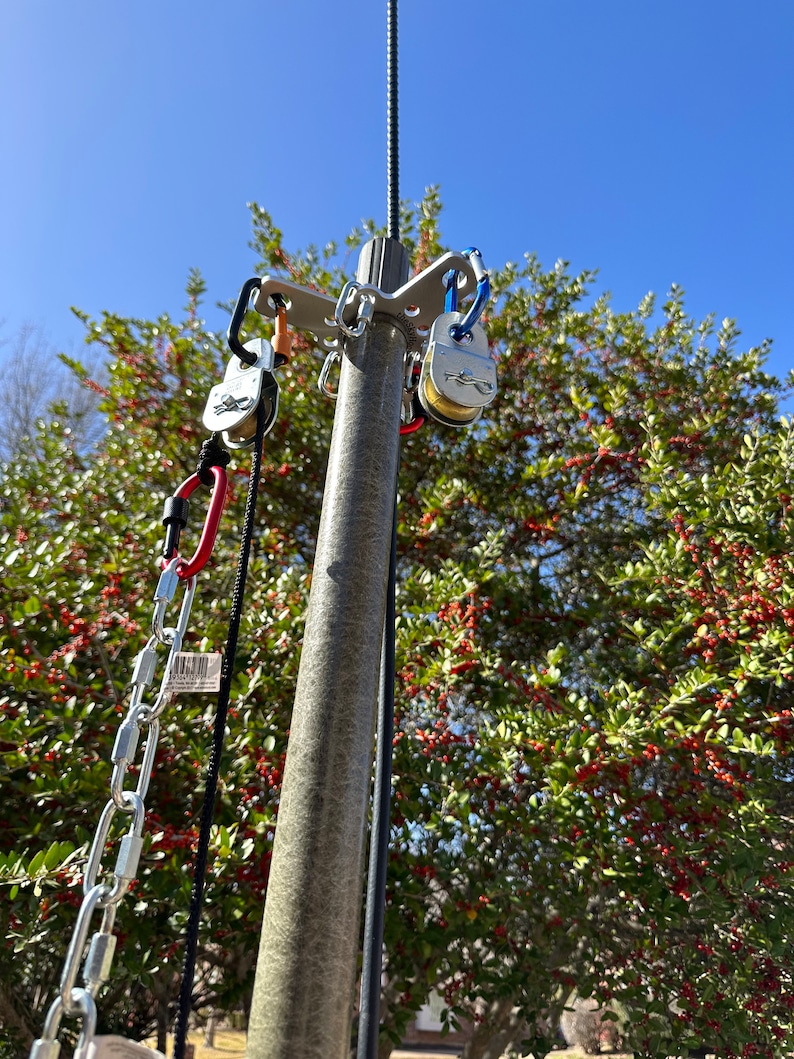 May include: A metal swing set attachment with multiple carabiners and pulleys. The attachment is mounted on a grey pole, with a chain and rope hanging down. The background features a clear blue sky and a tree with red berries.