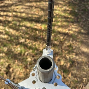 May include: A silver antenna base with a cylindrical support and a black, spiraled antenna. The base has a triangular shape with the text "theSkyH" and "ehamstop.com" printed on it. The antenna is set against a blurred outdoor background.