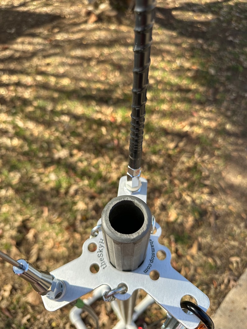 May include: A silver antenna base with a cylindrical support and a black, spiraled antenna. The base has a triangular shape with the text "theSkyH" and "ehamstop.com" printed on it. The antenna is set against a blurred outdoor background.
