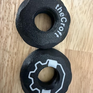 Op de afbeelding: Twee zwarte, cirkelvormige objecten met een getextureerd oppervlak. Eén heeft de tekst "theCroft" in het wit, de andere heeft een wit tandwielachtig ontwerp. De objecten liggen op een houten oppervlak.