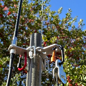 May include: A silver antenna mount with carabiners and a pulley system. A black cable and a silver antenna are also visible. The mount is attached to a grey pole, with green foliage and a blue sky in the background.
