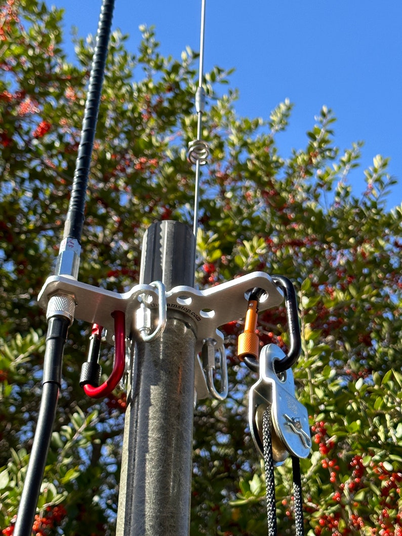 May include: A silver antenna mount with carabiners and a pulley system. A black cable and a silver antenna are also visible. The mount is attached to a grey pole, with green foliage and a blue sky in the background.