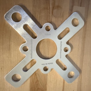 May include: A silver-colored metal bracket with an X-shaped design, featuring a central circular opening and multiple smaller holes. The text "theskydeck.com" and "Made in China" are visible. The bracket is on a wooden surface.