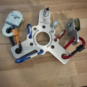 May include: A silver metal climbing device with multiple carabiners in blue, red, and black. The device has a central hub with the text "thehamstop.com" and "theSkyHook3x8". Two pulleys are attached to the device with orange and red accents.