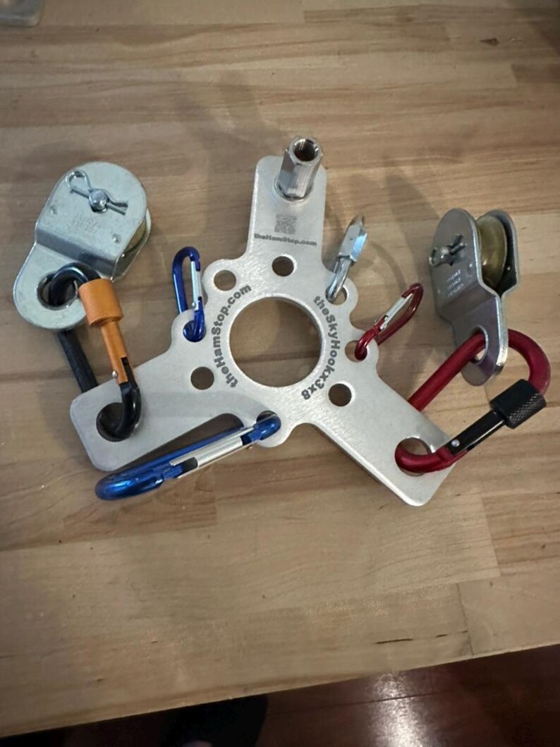 May include: A silver metal climbing device with multiple carabiners in blue, red, and black. The device has a central hub with the text "thehamstop.com" and "theSkyHook3x8". Two pulleys are attached to the device with orange and red accents.