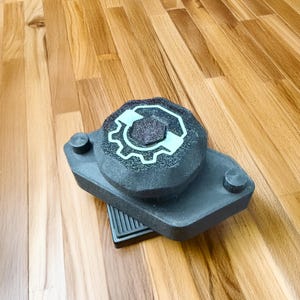 Puede incluir: Un objeto gris oscuro de estilo industrial con una parte superior circular con un diseño de engranaje en azul claro. El objeto tiene una base rectangular con dos pequeñas protuberancias cilíndricas y una sección acanalada.