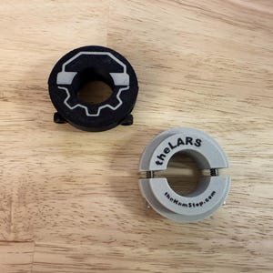 May include: Two circular, 3D-printed objects. One is black with a white gear and headphone design. The other is light gray, split with two metal springs, and has the text "theLARS" and "theHnmStop.com".