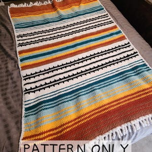 Pode incluir: Uma manta de crochê listrada colorida com uma borda de franjas. A manta é feita com uma variedade de cores, incluindo laranja, amarelo, azul e branco. A manta está colocada sobre uma colcha cinza. O texto "PATTERN ONLY" está impresso na imagem.