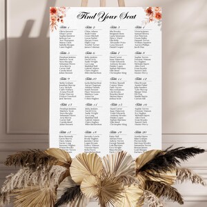 May include: A white seating chart with the words "Find Your Seat" in elegant script. The chart lists names under table numbers, with floral accents in the corners. The chart is displayed on an easel with a decorative floral arrangement.