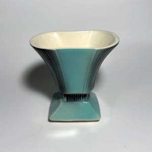 May include: A teal ceramic vase with a wide, flared, scalloped rim and a square base. The exterior features vertical ribbed detailing along the sides, while the interior is finished in a smooth, off-white glaze. The vessel stands upright.