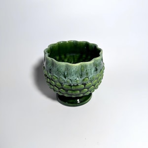 May include: A small, round, green ceramic bowl with a pedestal base. The exterior features a raised, textured, scale-like pattern. The upper rim is scalloped and transitions into a lighter, mottled green glaze that contrasts with the darker base.