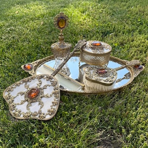 May include: A vintage vanity set with a mirrored tray, perfume bottle, hand mirror, brush, comb, and a small trinket box. The set features ornate gold-toned metalwork, amber-colored stones, and a white, floral-patterned compact.