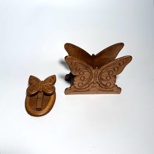 May include: A medium brown wooden napkin holder shaped like a butterfly with carved wing details, accompanied by a matching small wooden butterfly clip on an oval base. Both items feature a smooth, polished finish and intricate scrollwork.
