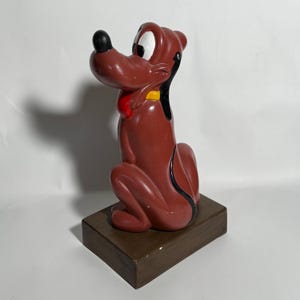 May include: A brown Pluto figurine with a black nose, white eyes, and a red collar. The figurine is sitting on a brown rectangular base. The dog is in a seated position, with its ears perked up.