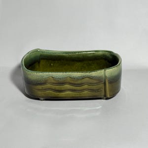 May include: A rectangular ceramic planter with a wavy design on the side. The planter is glazed in shades of green, with a darker interior and lighter, textured edges. It has small feet and a smooth, glossy finish.