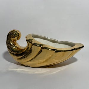 May include: A gold-colored, shell-shaped decorative planter. The planter has a glossy finish and a white interior. The design features a curled accent on one end. The planter is suitable for displaying small plants or decorative items.