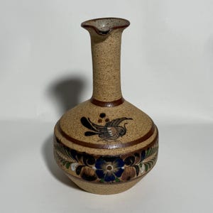 May include: A tan ceramic vase with a rounded body and a tall, slender neck. The vase features a bird design and floral patterns in brown, blue, and green. The vase has a spout and is approximately 8 inches tall.