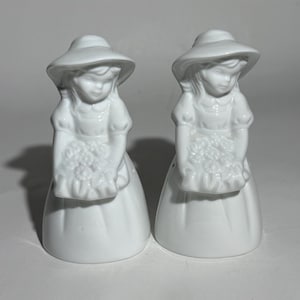 May include: Two white ceramic figurines of girls wearing hats and dresses, each holding a bouquet of flowers. The figurines have a classic, vintage design, with detailed facial features and clothing. The salt and pepper shakers are a charming addition to any kitchen or dining room.