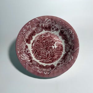May include: A round ceramic bowl featuring a deep maroon transferware pattern. The center depicts a horse-drawn carriage in a rural landscape, while the wide rim is decorated with a dense floral motif of large blossoms and leaves in matching maroon.