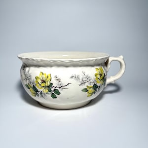 May include: White ceramic bowl with a handle and a scalloped rim. The bowl is decorated with yellow floral and green leaf designs. The handle is curved and has a decorative design. The bowl is likely vintage.