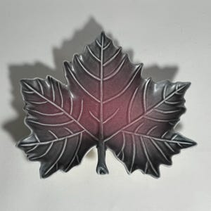 May include: A gray and pink ceramic leaf-shaped dish. The dish is shaped like a maple leaf and has a raised design.