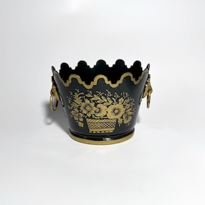 May include: Black metal decorative bucket with gold floral design and lion head handles. The bucket has a scalloped rim and a gold trim around the top and base. The floral design features flowers in a basket.