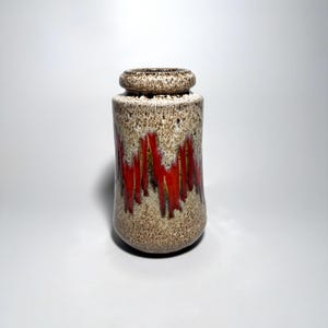 May include: A ceramic vase with a cylindrical body and a rounded rim. The surface features a speckled beige and brown base glaze with a prominent vertical drip pattern in bright red that cascades down the center of the vessel's exterior.