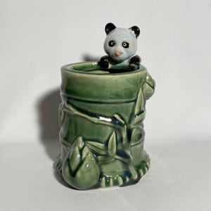 May include: A ceramic panda bear figurine peeking over the rim of a green bamboo-style container. The panda has black and white markings. The container is a glossy green with bamboo stalk and leaf detailing.
