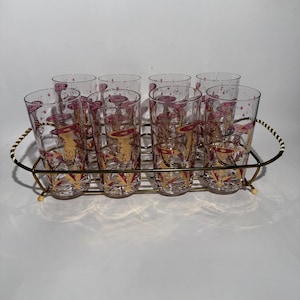 May include: A set of eight vintage highball glasses with a gold and pink design, displayed on a gold-toned metal serving tray. The glasses feature a repeating pattern of stylized shapes and starbursts. The tray has a decorative handle.
