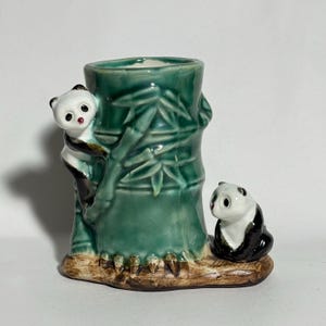 May include: A ceramic panda bear figurine with a green bamboo-shaped vase. One panda is climbing the bamboo, and another is seated at the base. The vase and pandas are painted in black, white, and green.