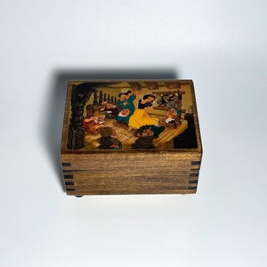 May include: A wooden box with a colorful illustration of Snow White and the Seven Dwarfs dancing. The box has a dark wood grain finish and is rectangular with small feet.