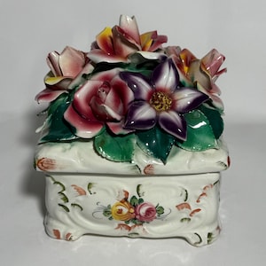 May include: A decorative ceramic box with a floral arrangement on top. The flowers include pink, red, and purple roses and other blooms, with green leaves. The box is white with painted floral designs.